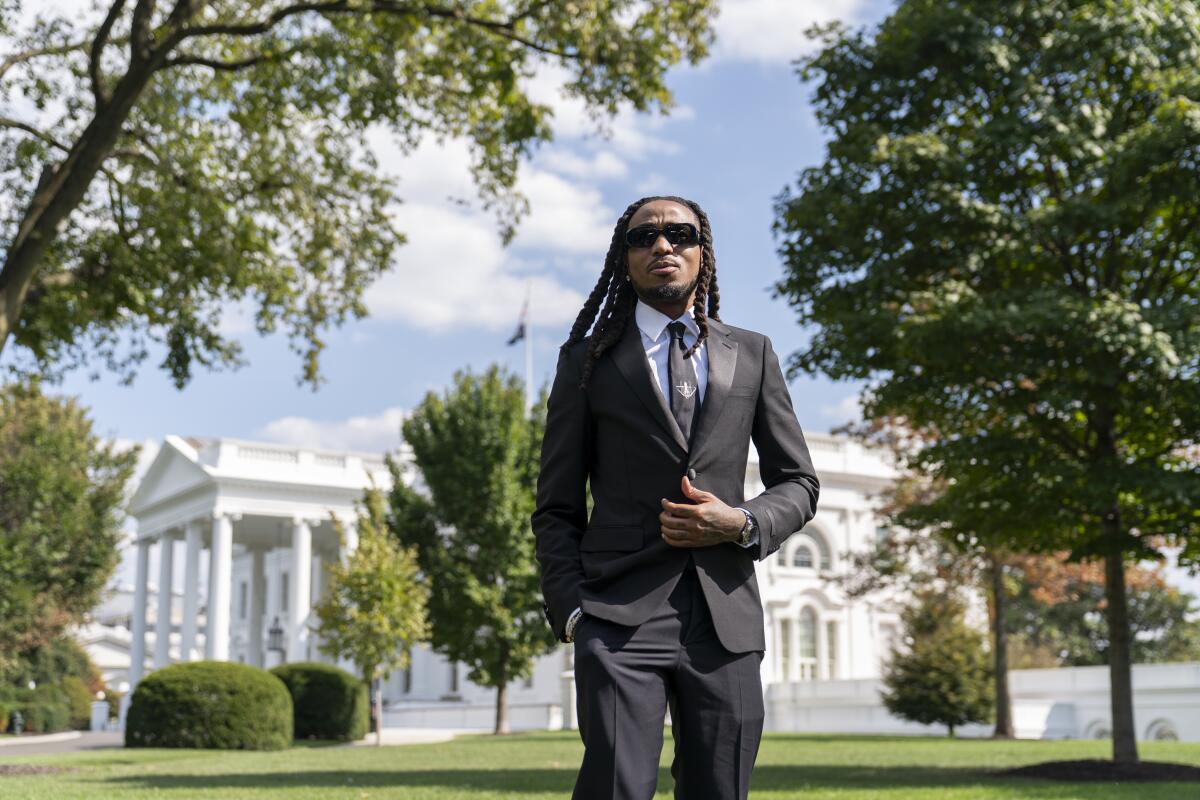 Quavo holds his jacket buttons with one hand as he stands in a dark suit and tie outside the White House.
