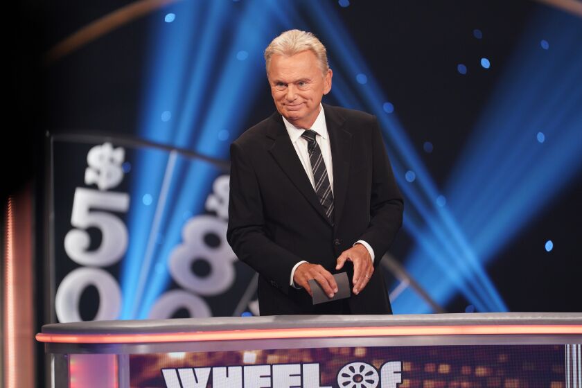 Man Pat Sajak standing on Wheel of Fortune stage in black suit, holding cards, smiling and looking off stage