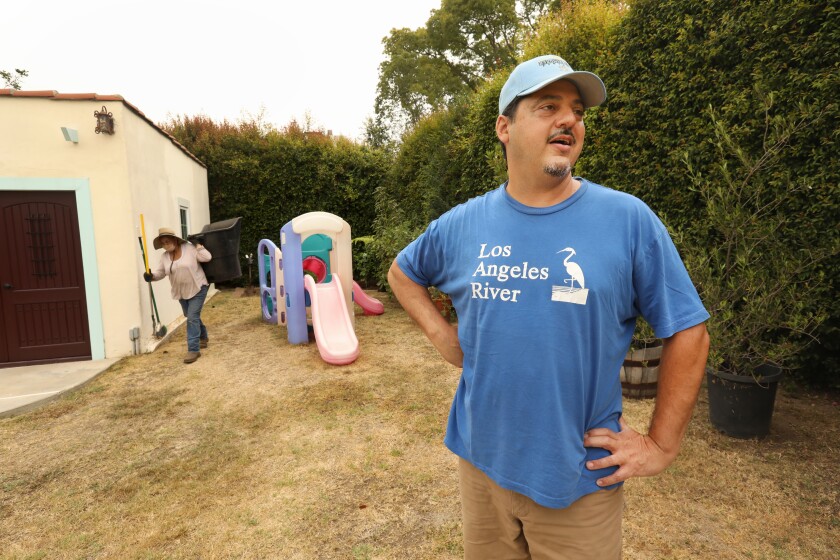 A man in a blue T-shirt and cap stands on a brown lawn.  Behind him, a woman in a hat holds a rake, a broom and a dark bin.  