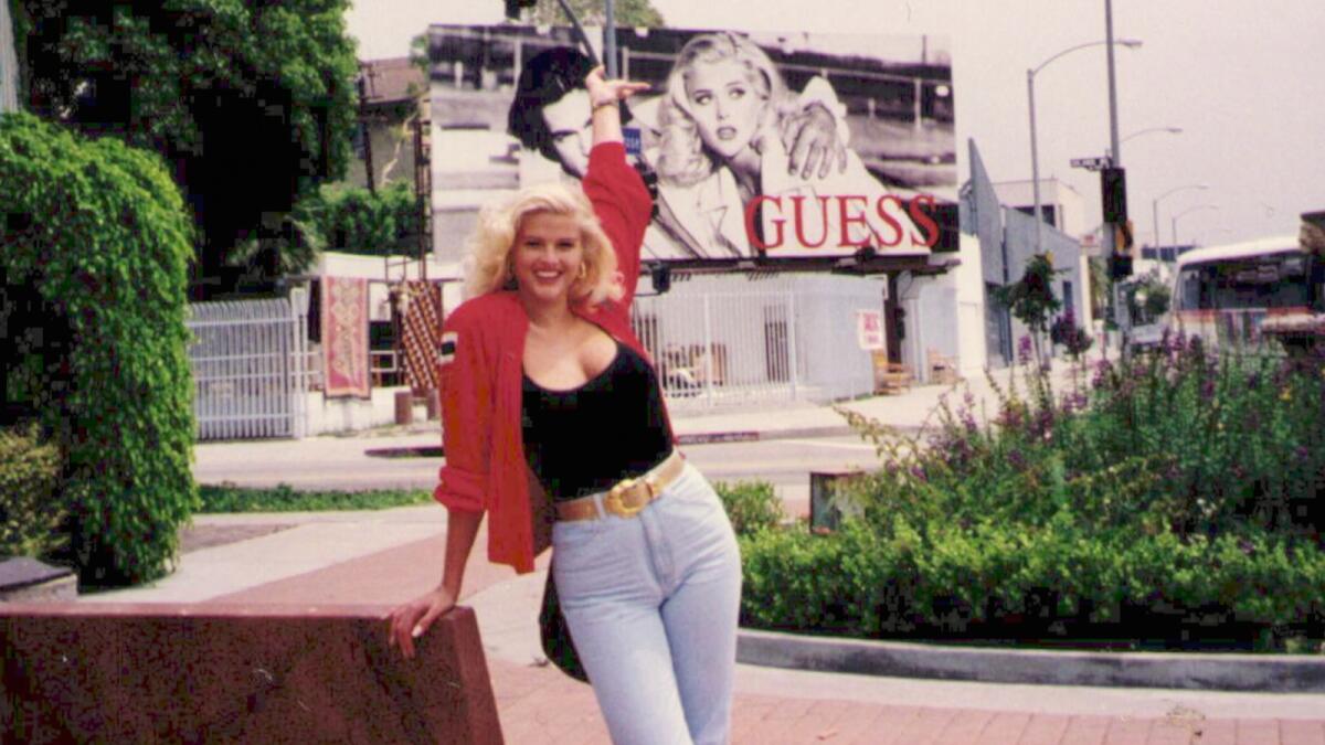 A smiling Anna Nicole Smith leans on a wall with one arm lifted in the air