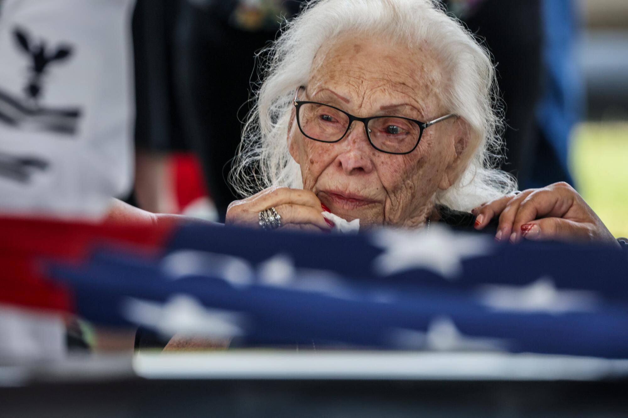 A woman with white hair and glasses near a U.S. flag