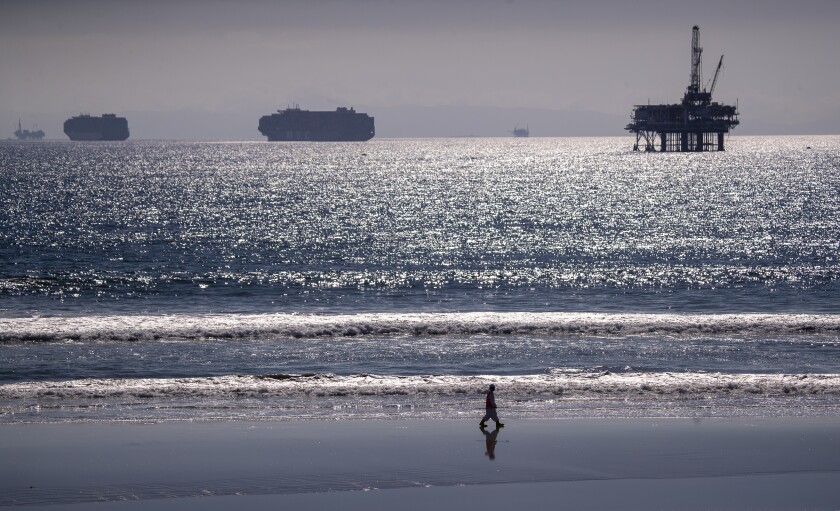 A person walks along the beach as container ships and an offshore oil rig loom in the background.
