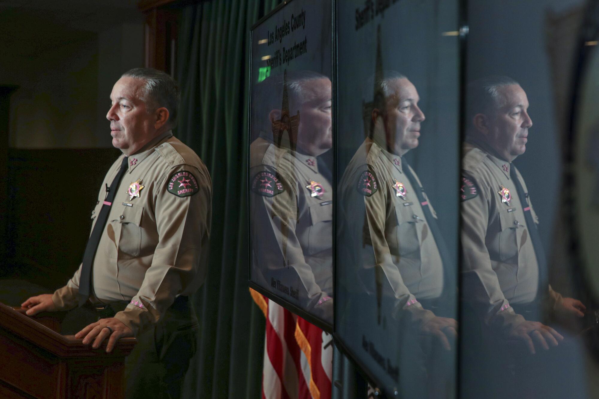 Sheriff Alex Villanueva is reflected on three monitors while speaking at a lectern