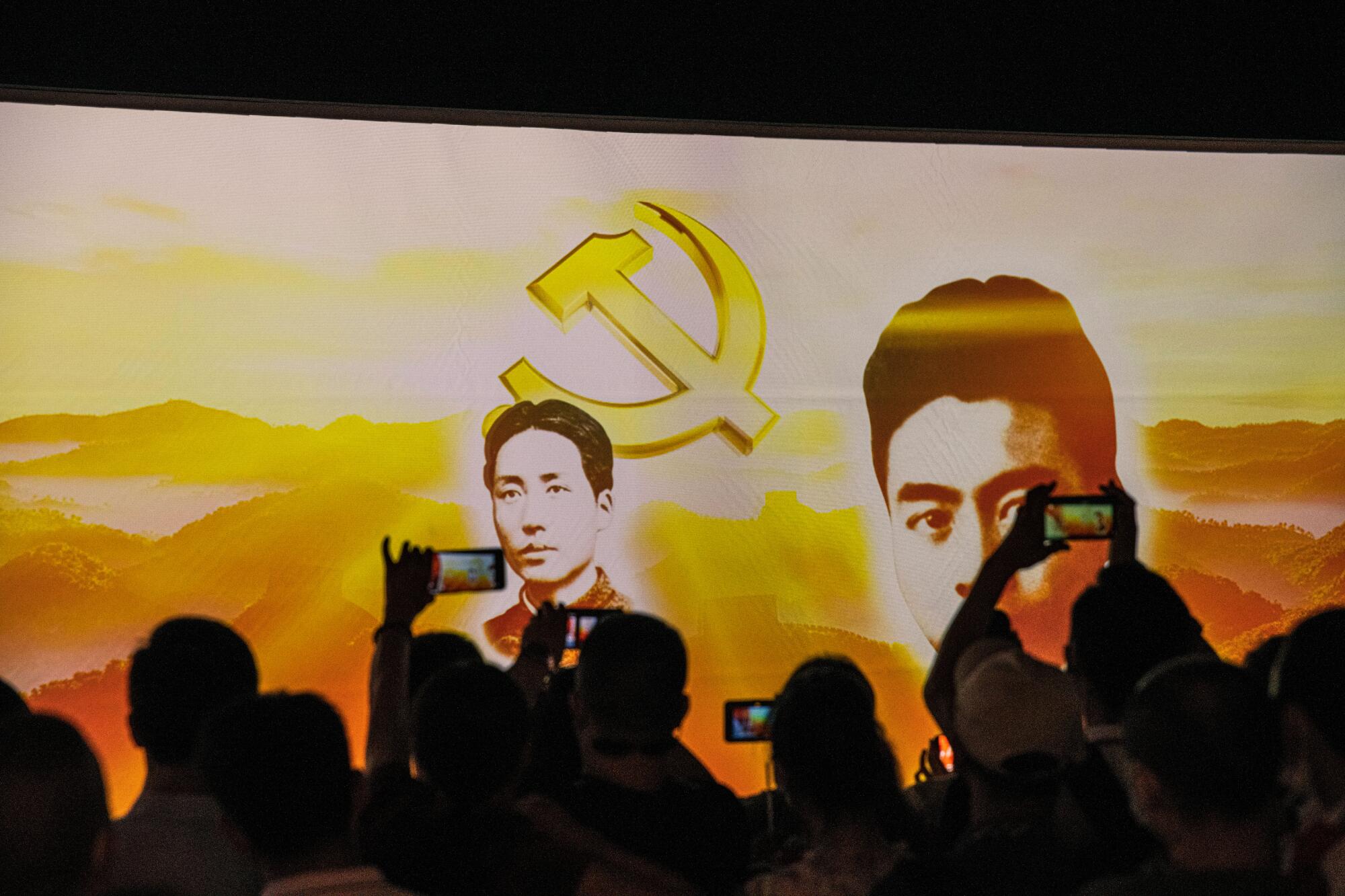 People hold up cellphones to record images of two men on screen, with a hammer and sickle symbol in the background 