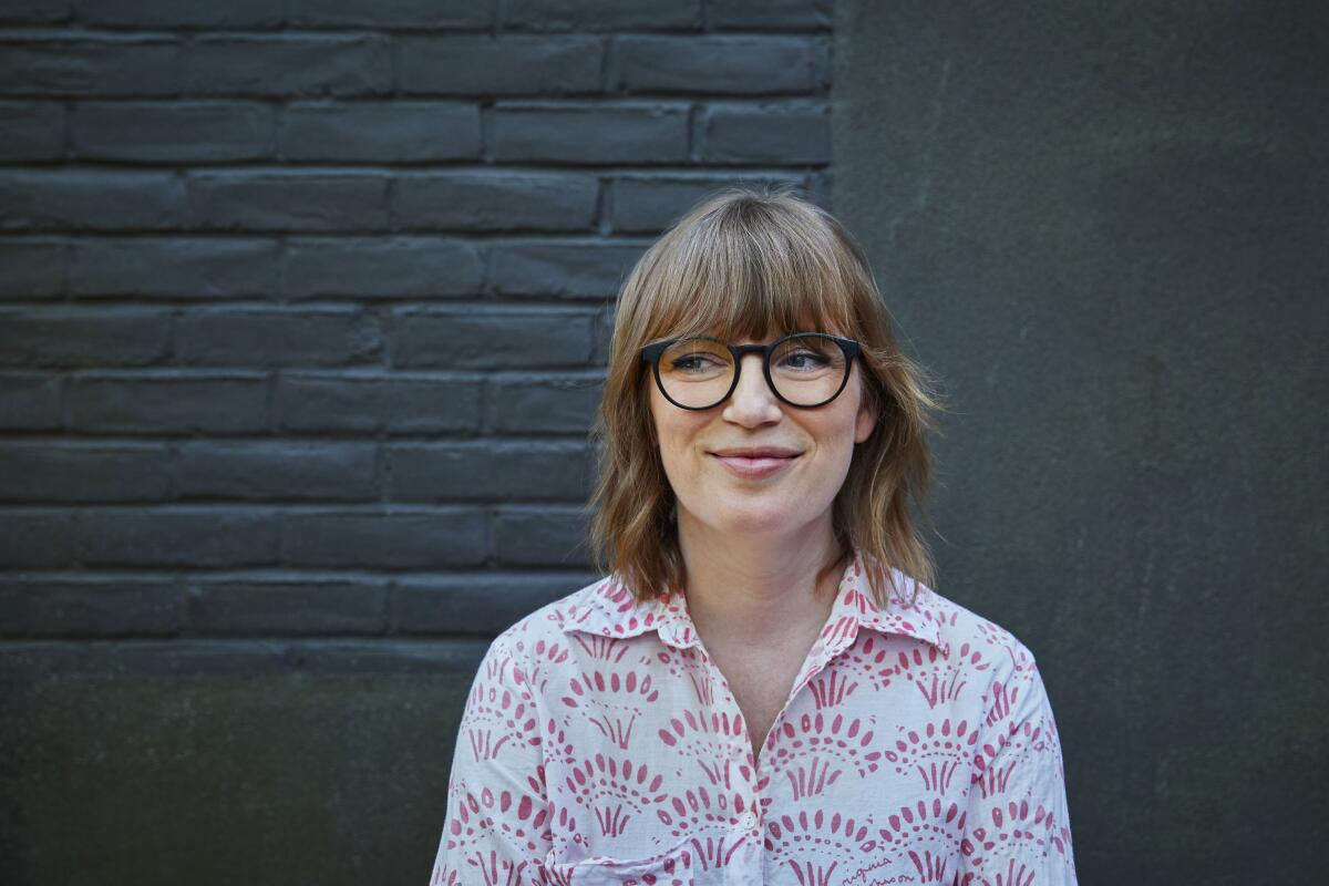 A woman with bangs, glasses and a button-down shirt stands against a gray brick wall.