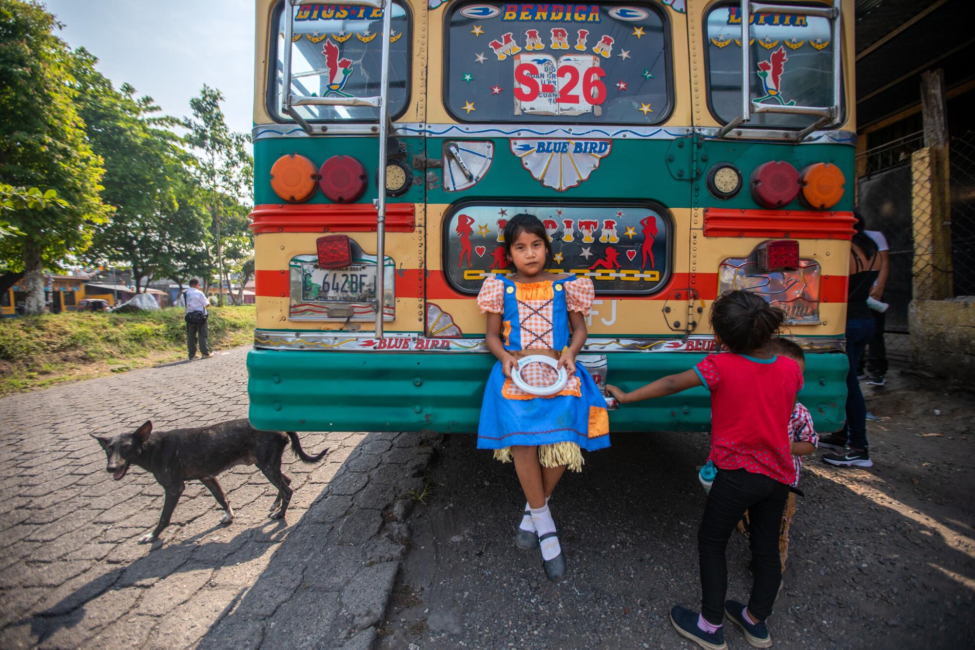 A girl leans sits on the back bumper of a colorful bus.