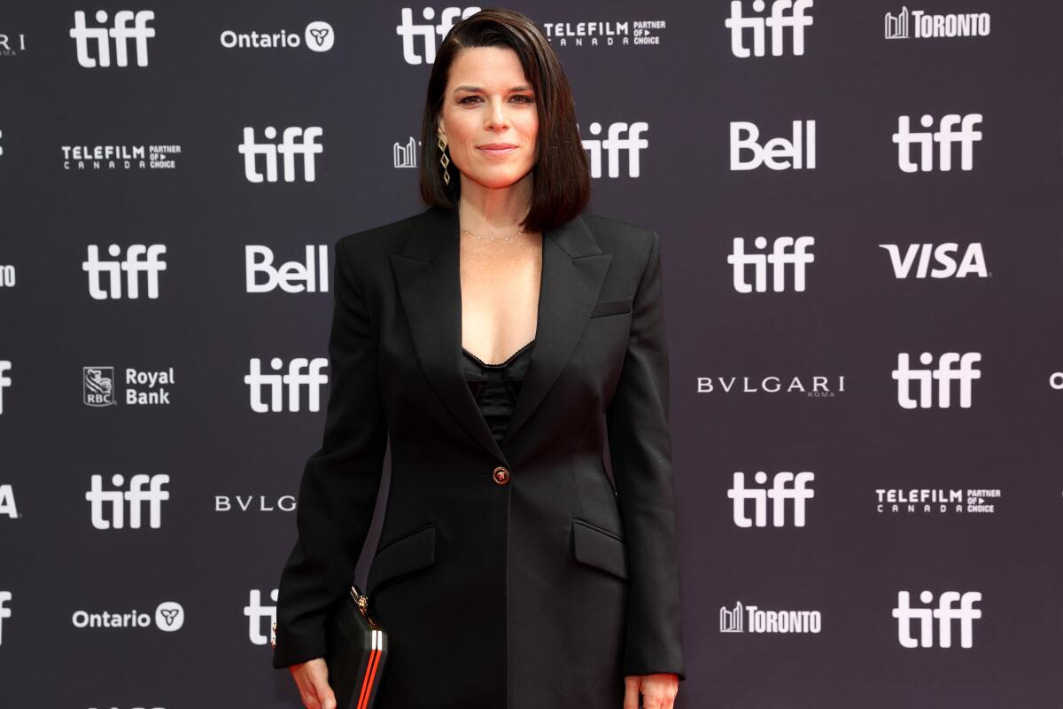 Neve Campbell poses for a photo in a fitted black suit jacket and top with plunging neckline.