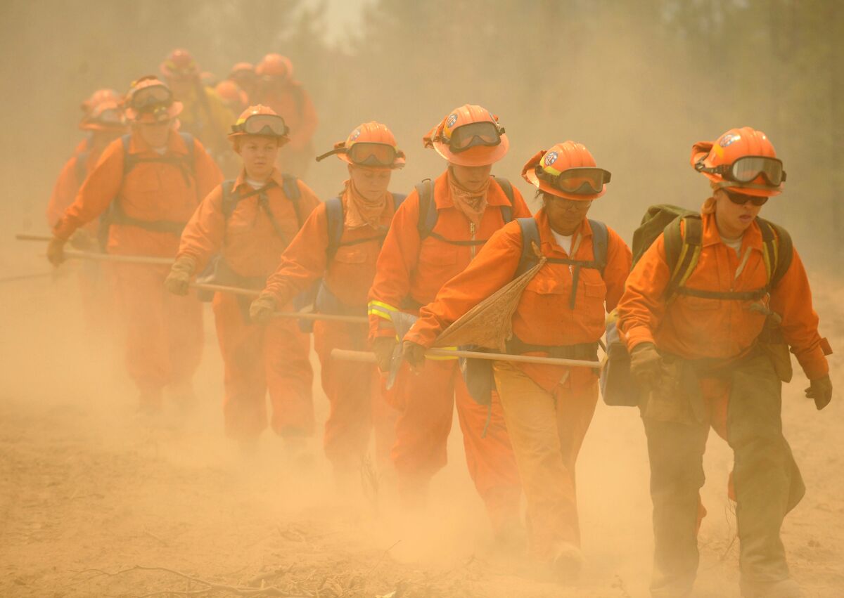 Women wearing orange fire suits and holding heavy equipment walk single file through smoke 
