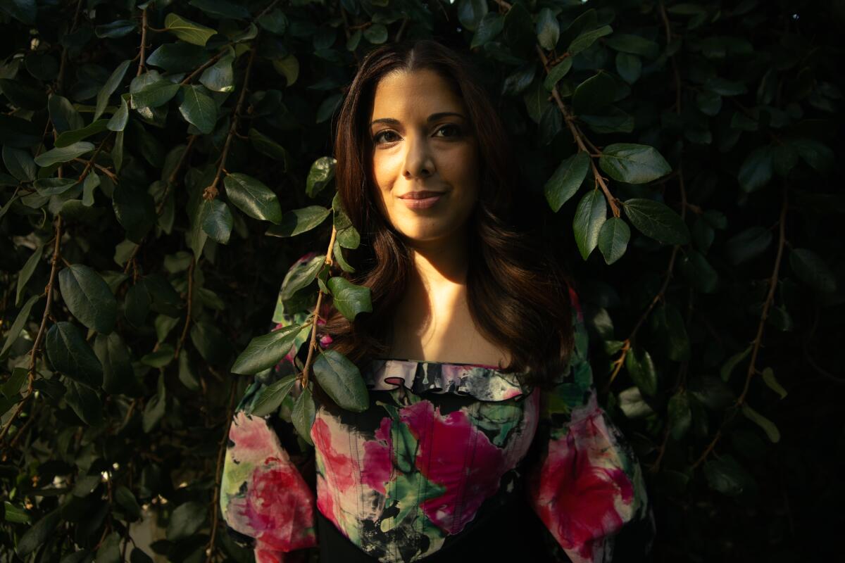 A woman in a floral blouse stands outside in front of dark green foliage.