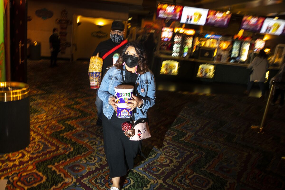 A man and a woman wearing masks carry popcorn and drinks through a lobby