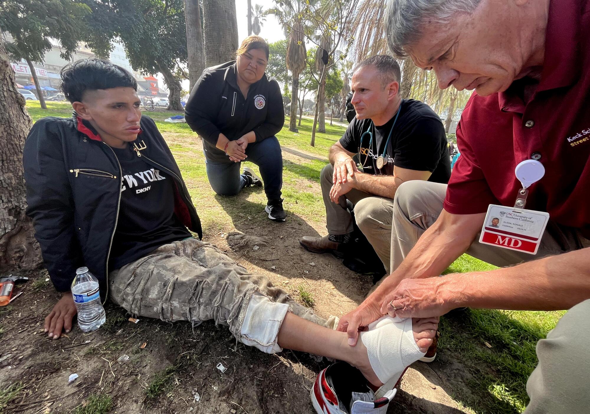 A man treats another's injured foot outdoors in a park. 