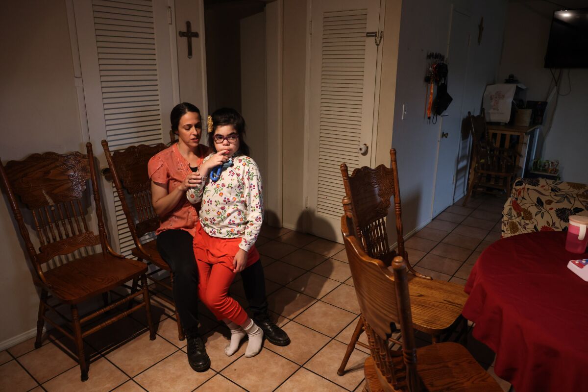 A 9-year-old girl with Sanfilippo syndrome, a degenerative disease, leans against her mother in their kitchen.