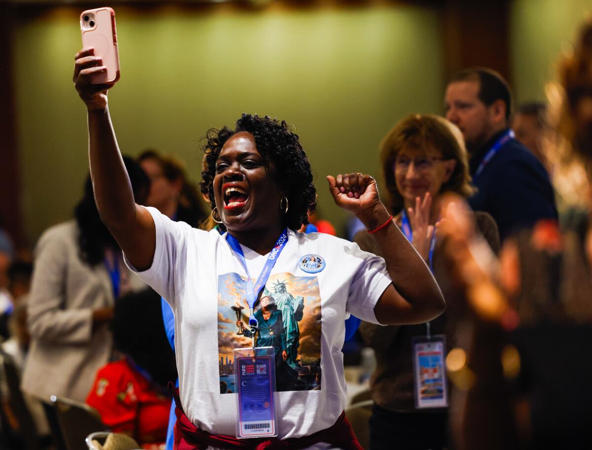 A woman with dark hair, wearing a white T-shirt, smiles while holding up a phone as other people applaud behind her