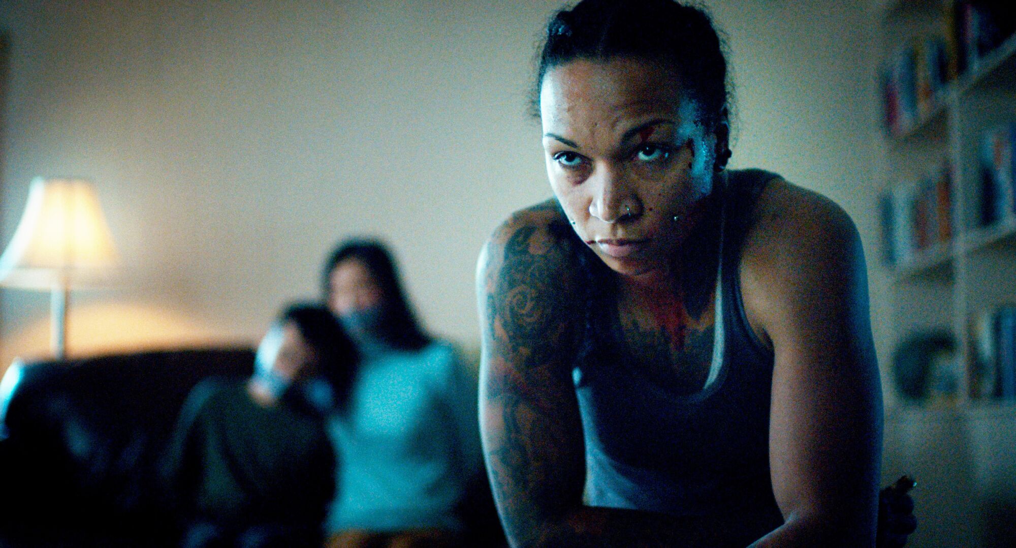 A closeup of a tattooed woman staring intently at the camera, with two people out of focus seated behind her.