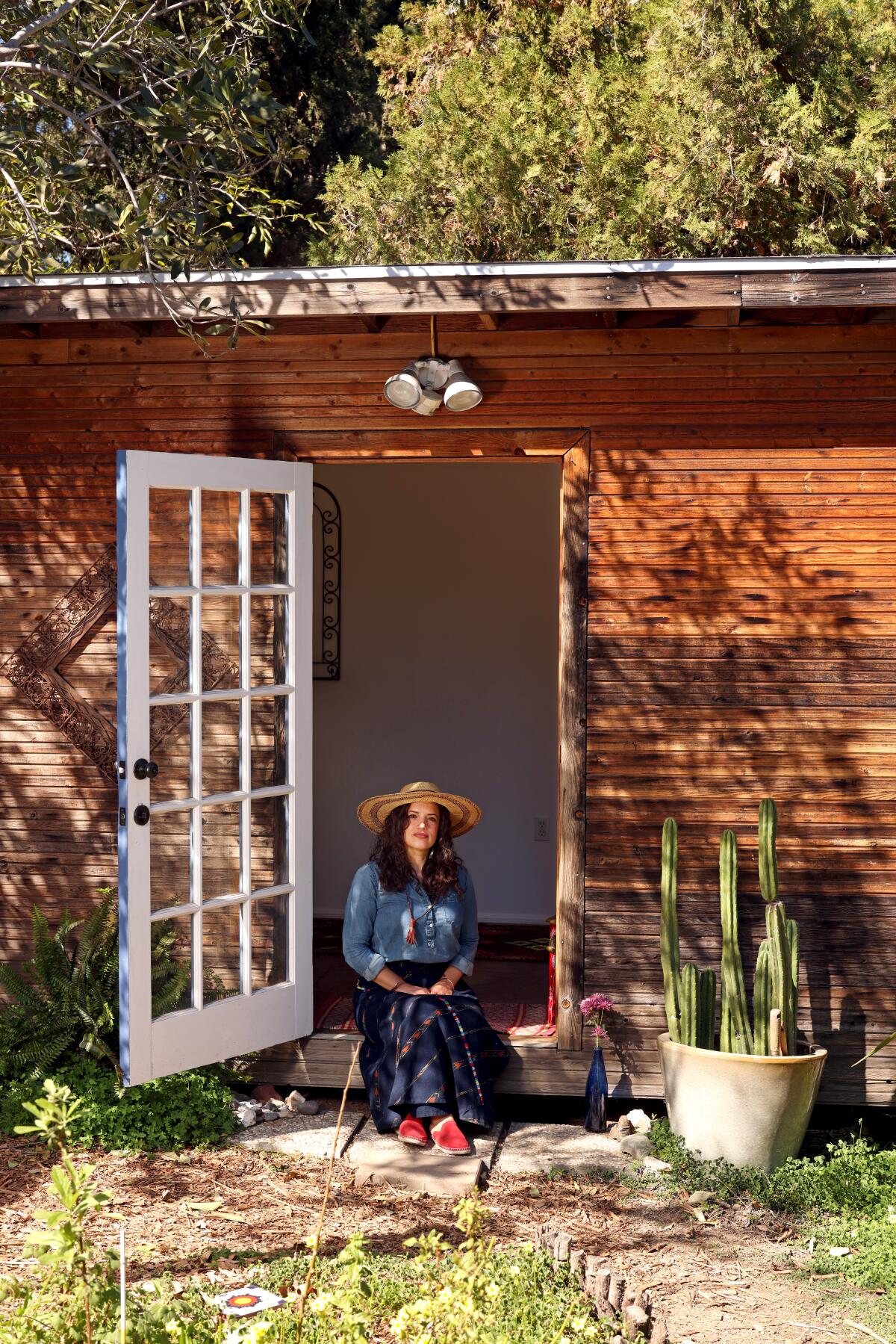 A woman wearing a hat smiles while sitting in the doorway of a wood structure.