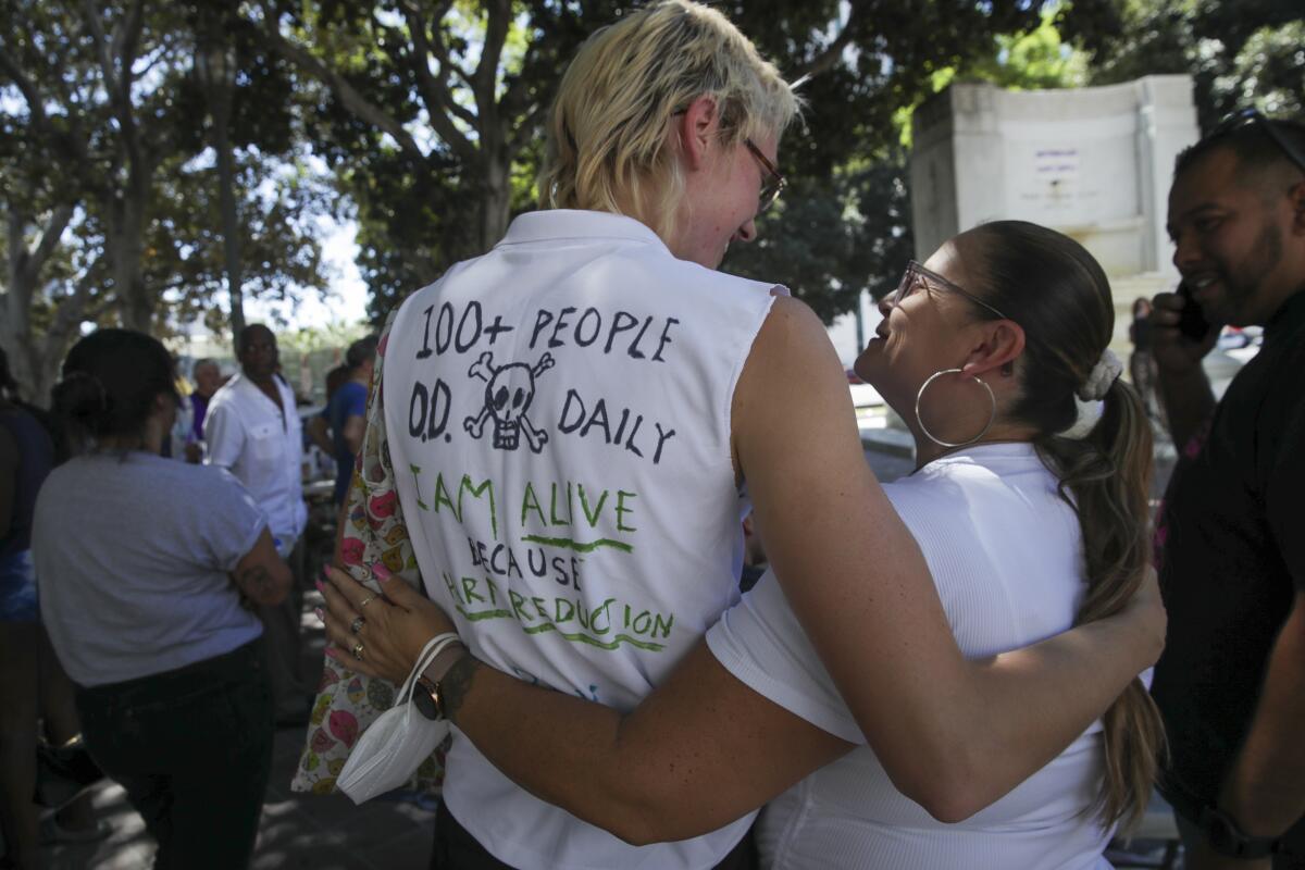 Two people hug, one wearing a shirt that says "100+ people O.D. daily"