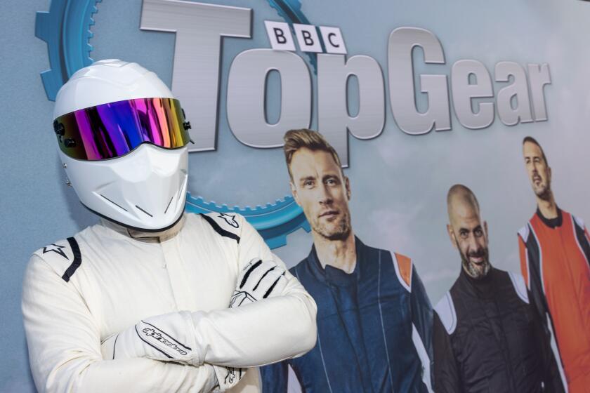 A racer in a white driving suit and helmet posing against a poster for "Top Gear" that shows three men standing and smiling