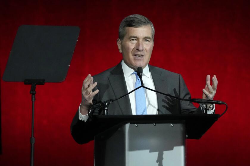 Charles Rivkin gestures at a podium while speaking into a microphone and wearing a gray suit with a blue tie.