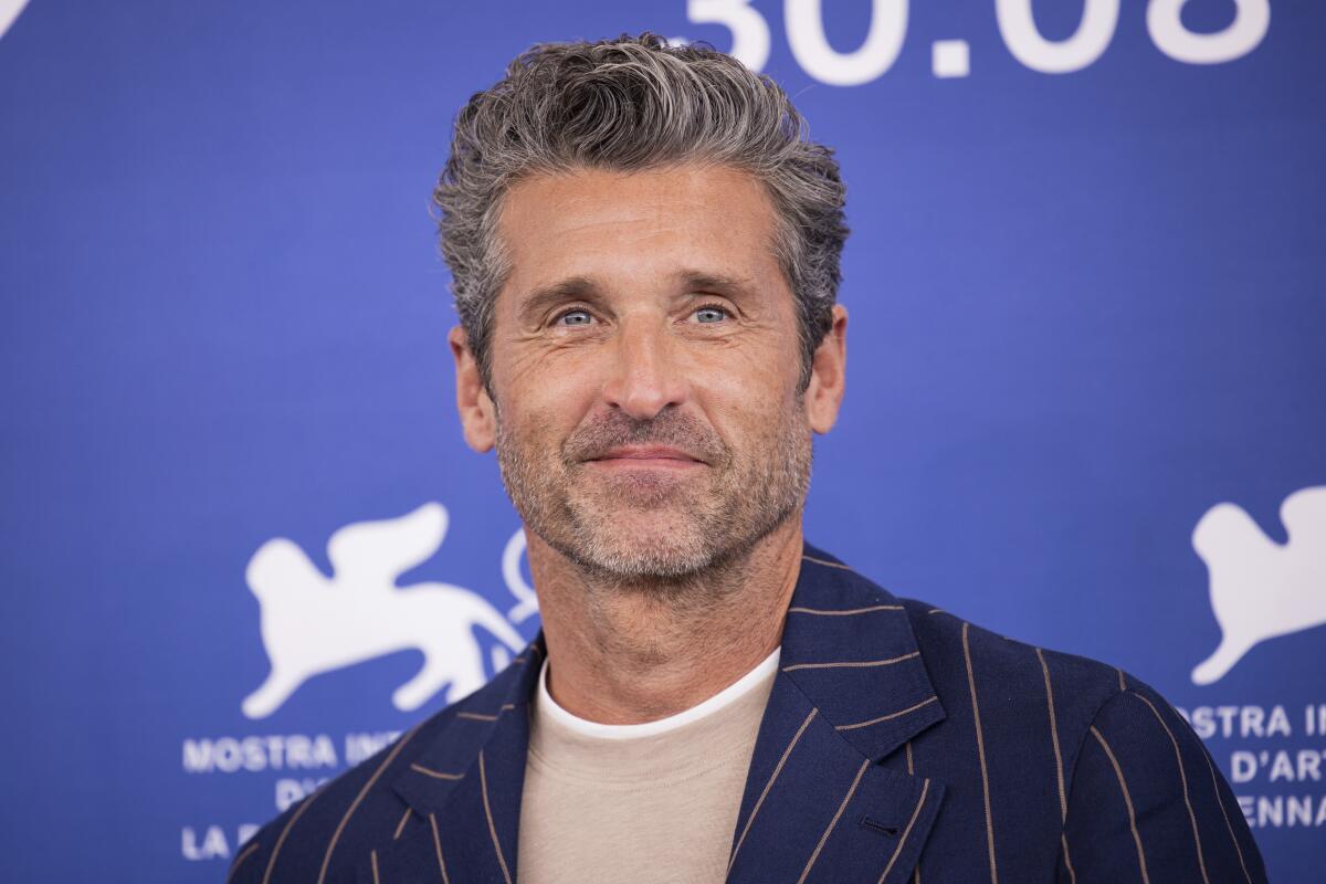 Patrick Dempsey with graying hair smiling in a tan shirt and a striped suit jacket against a blue backdrop