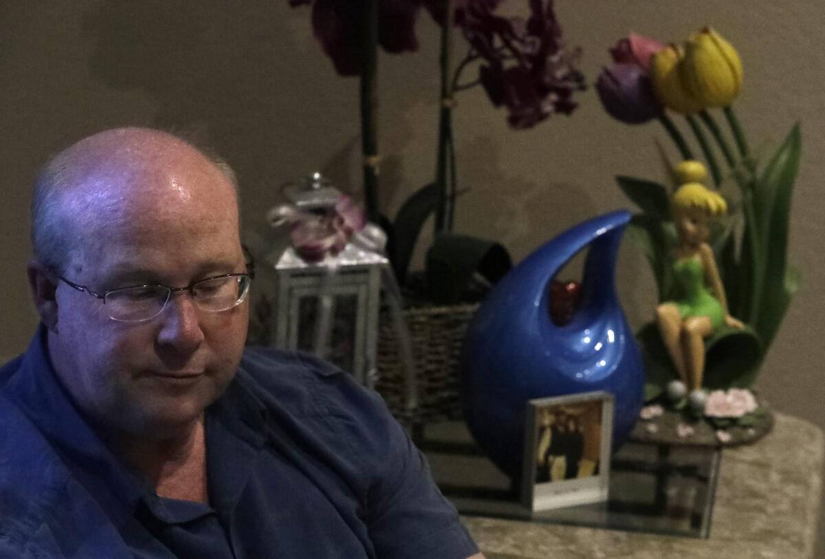 A man with glasses sits before a table of flowers, and urn and mementos