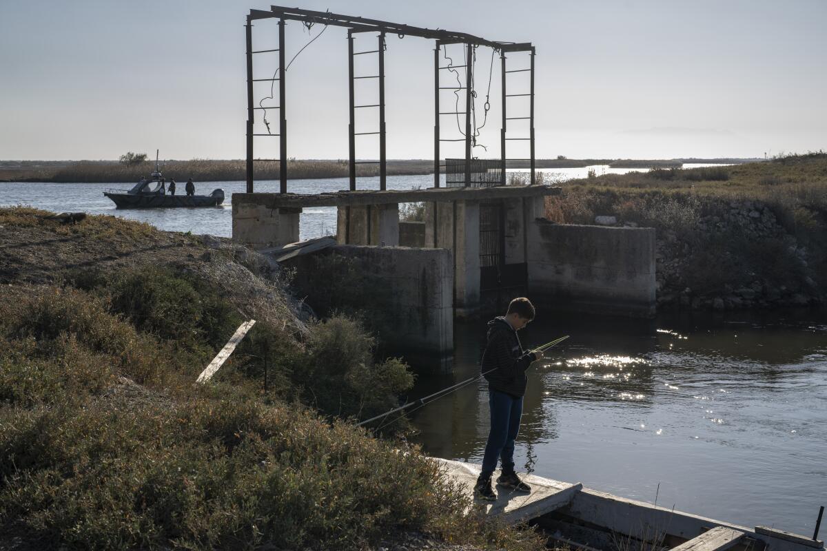 A boy fishes as, in the background, police border patrol boats sail along the Evros River. 