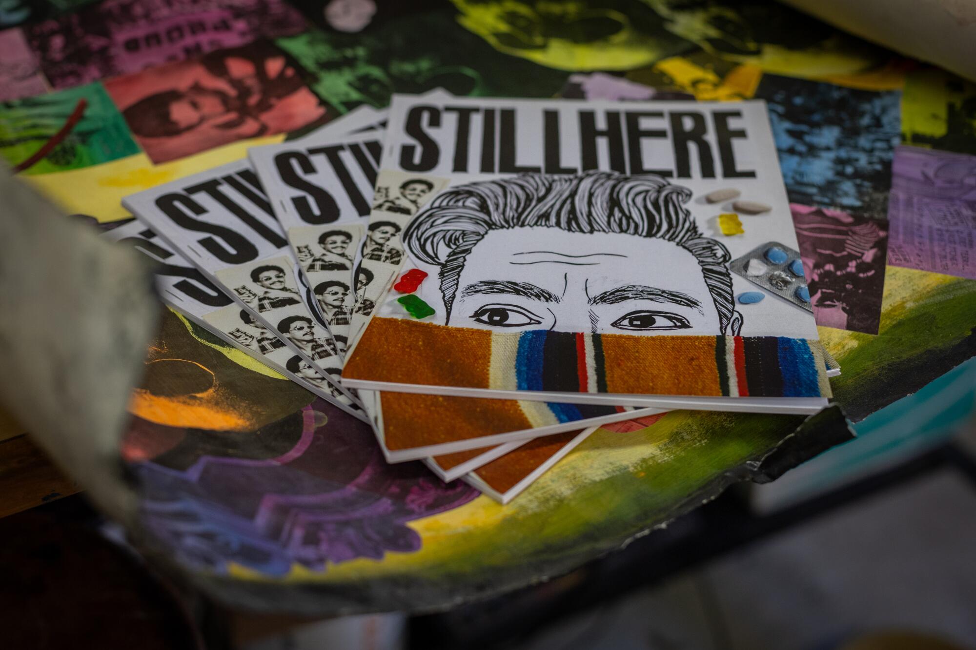 A stack of zines titled "STILL HERE" show a drawing of the top of a man's face obscured by a Mexican serape.