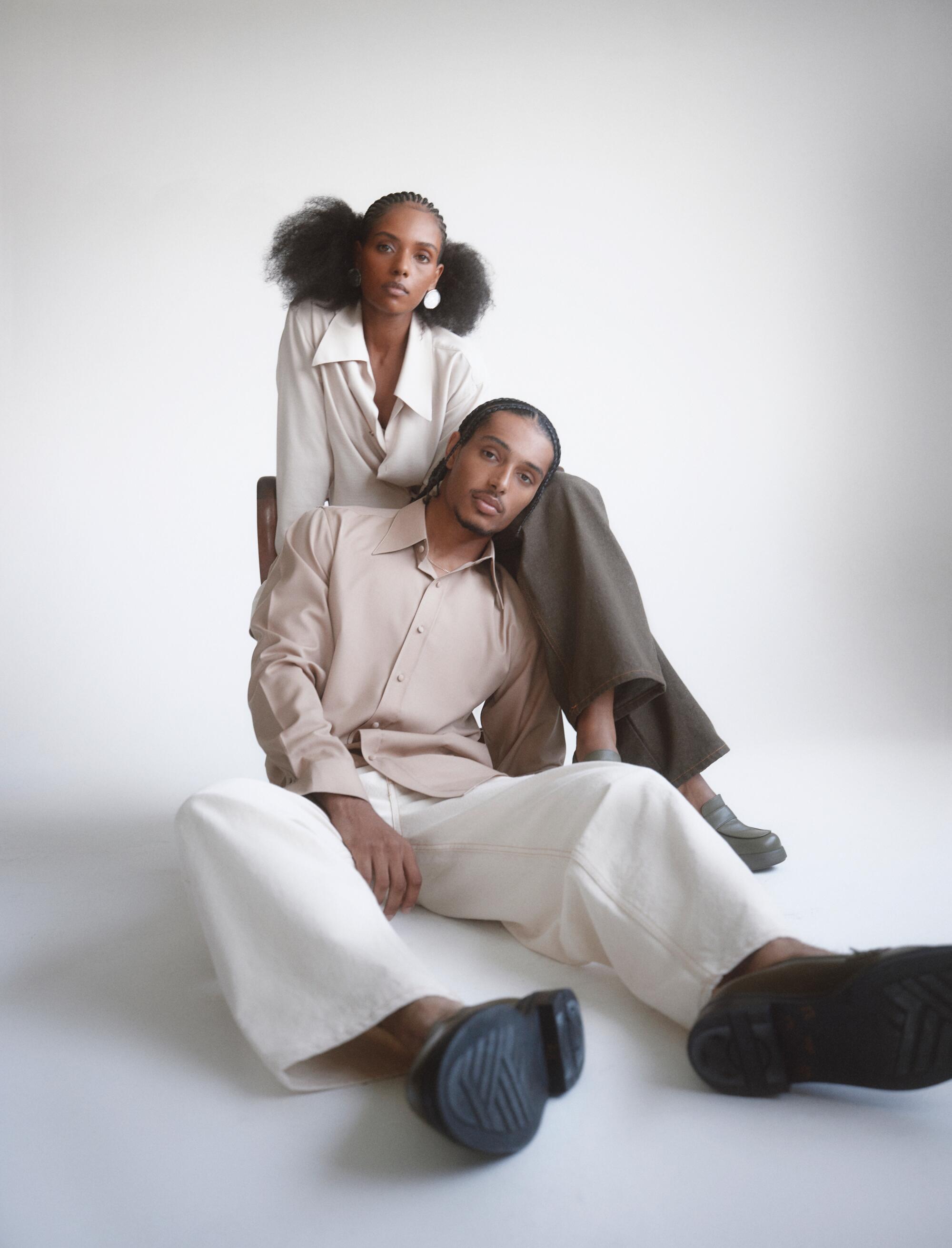 A male model sits on the floor, leaning his head on the leg of a female model. Both wear cream-colored outfits.