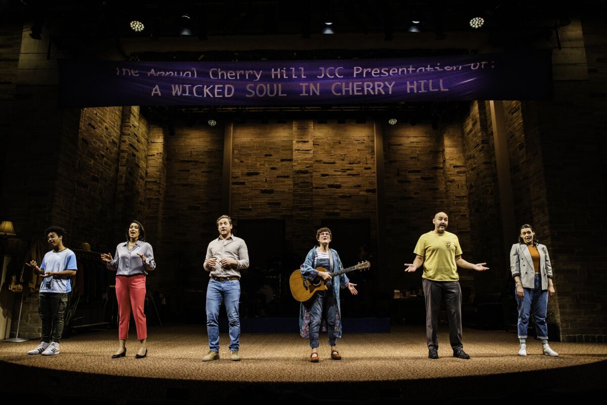 Six people, one holding a guitar, stand in line on a stage.