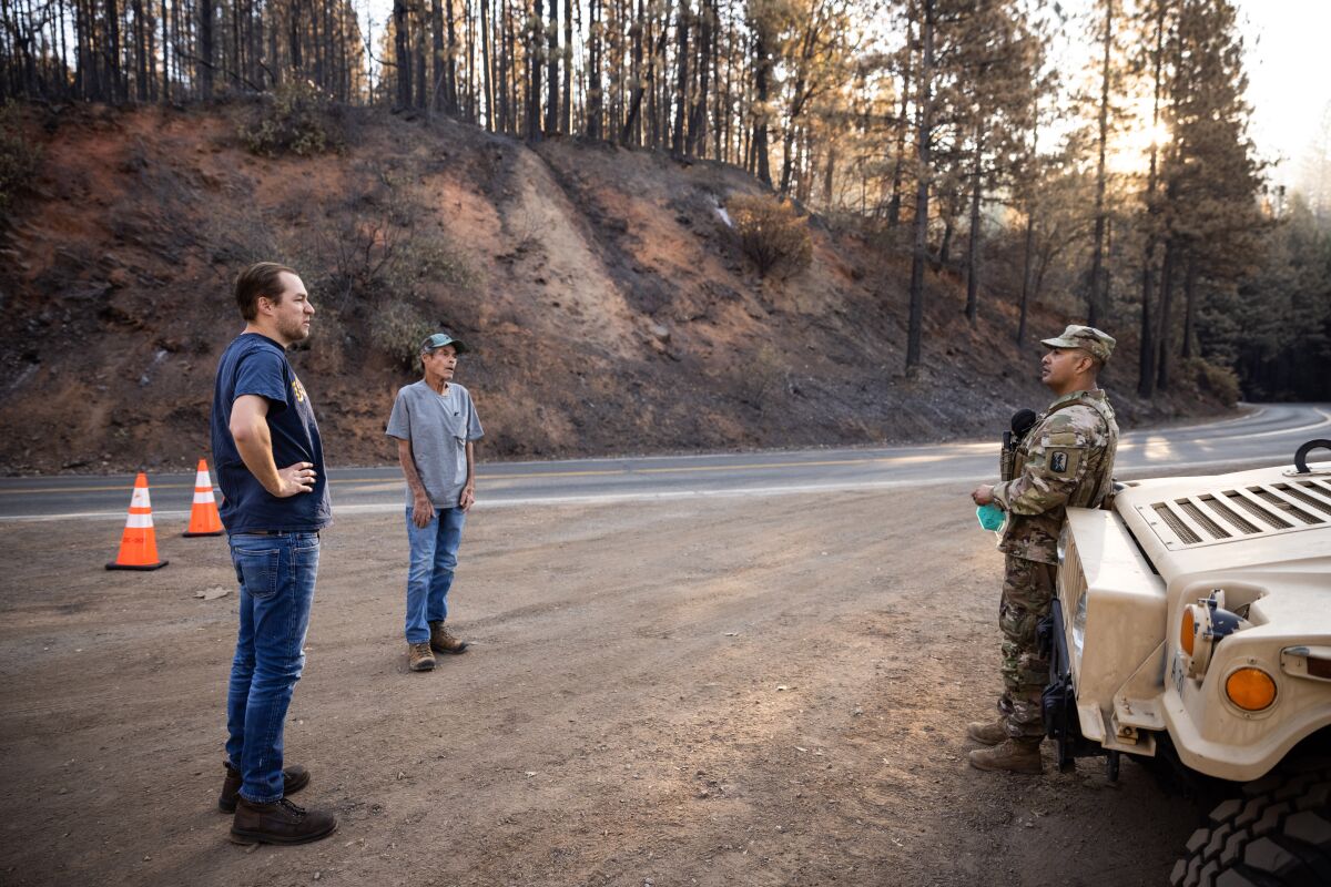 Two men in jeans talk to a man in military uniform by a road.