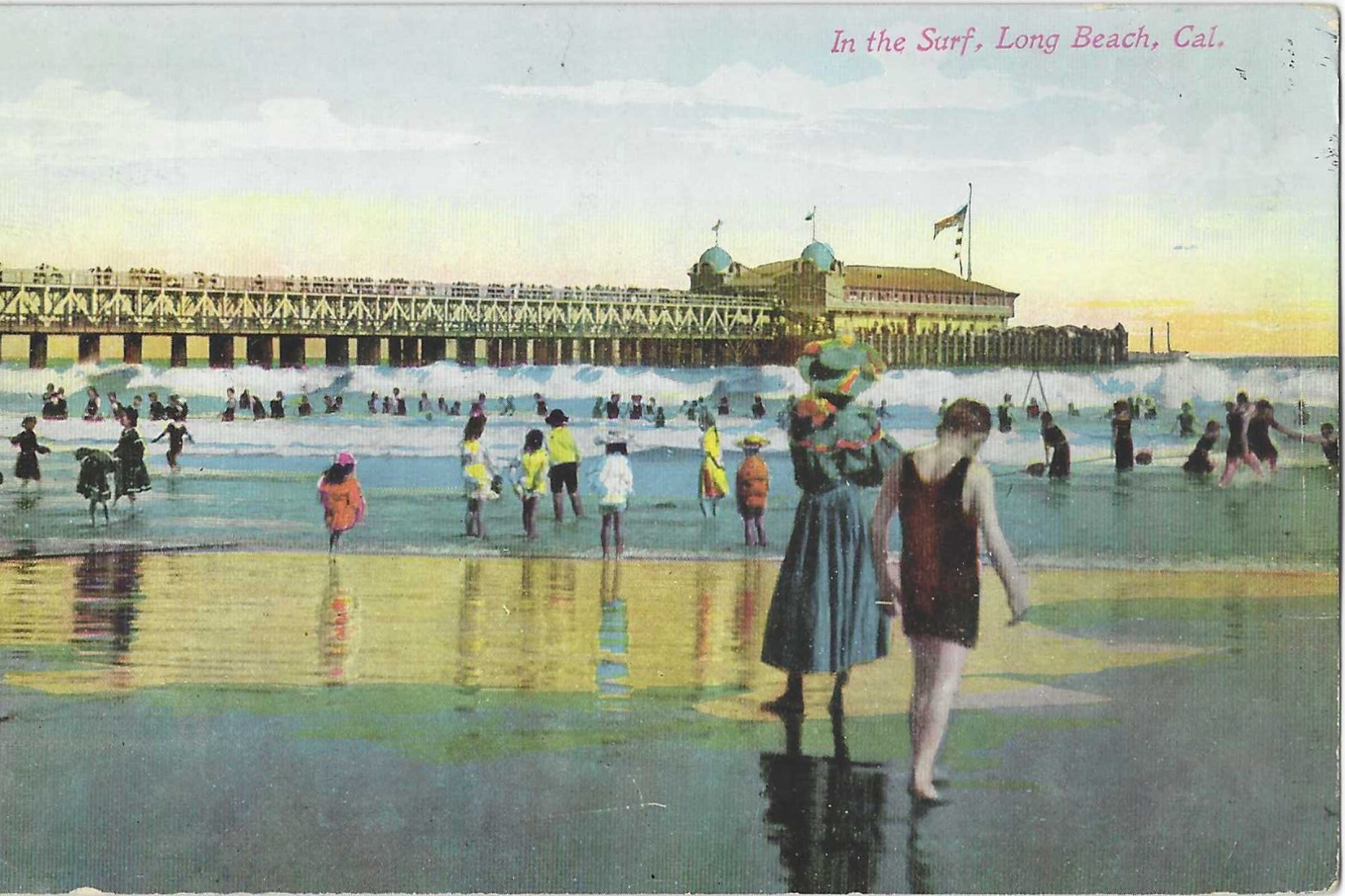 People play in the surf near a pier. Prominent in the frame is a woman in an ankle-length blue dress and a hat