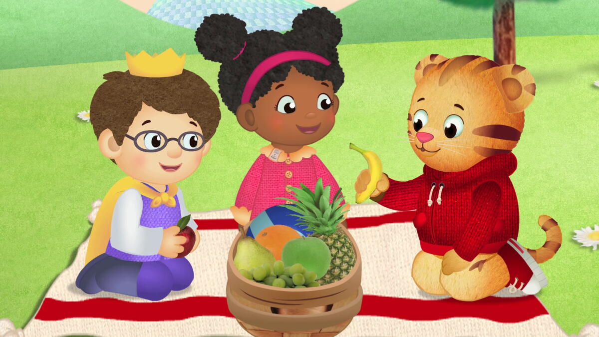 An animated still of a little boy, girl and a tiger sitting around a bowl of fruit on a picnic blanket.