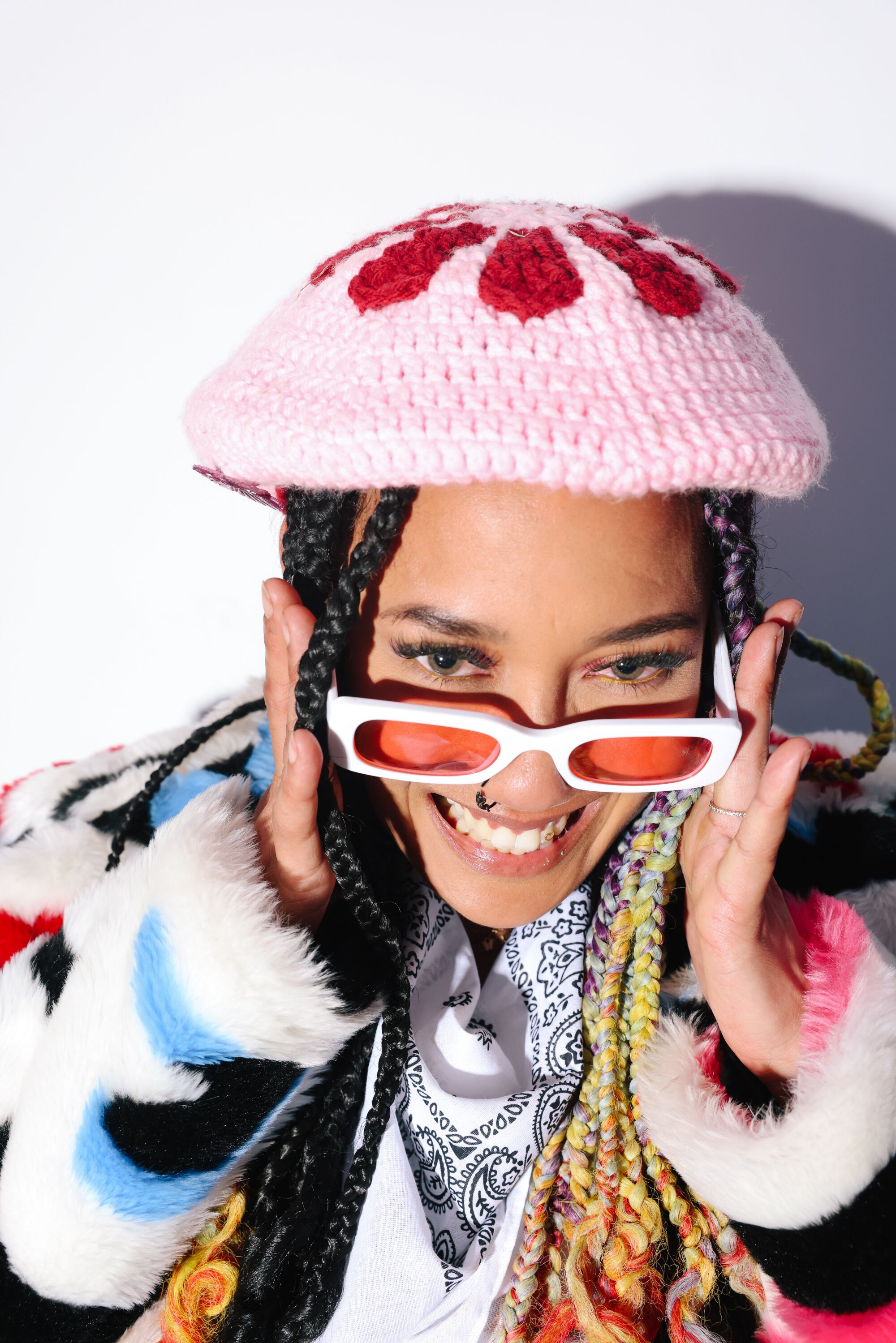 A woman poses wearing a colorful furry rainbow coat, pink and red crocheted hat and white sunglasses with pink lenses