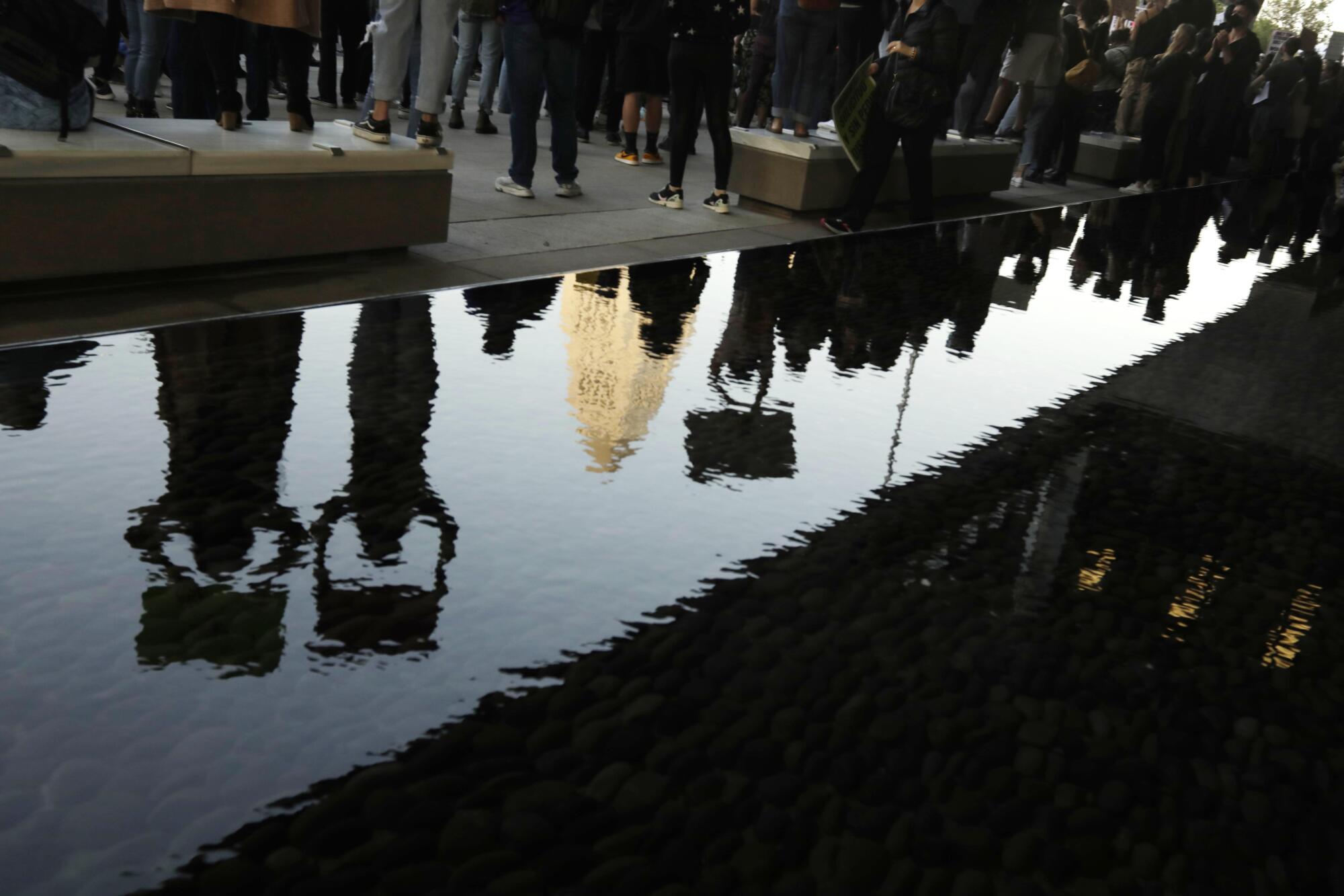 A crowd of people, some holding signs, are reflected in water