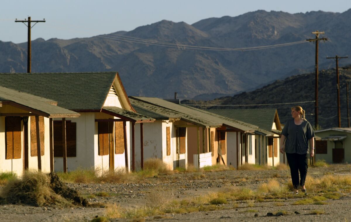 A woman at right walks past a row of boarded-up single-story homes. A mountain range is on the horizon. 