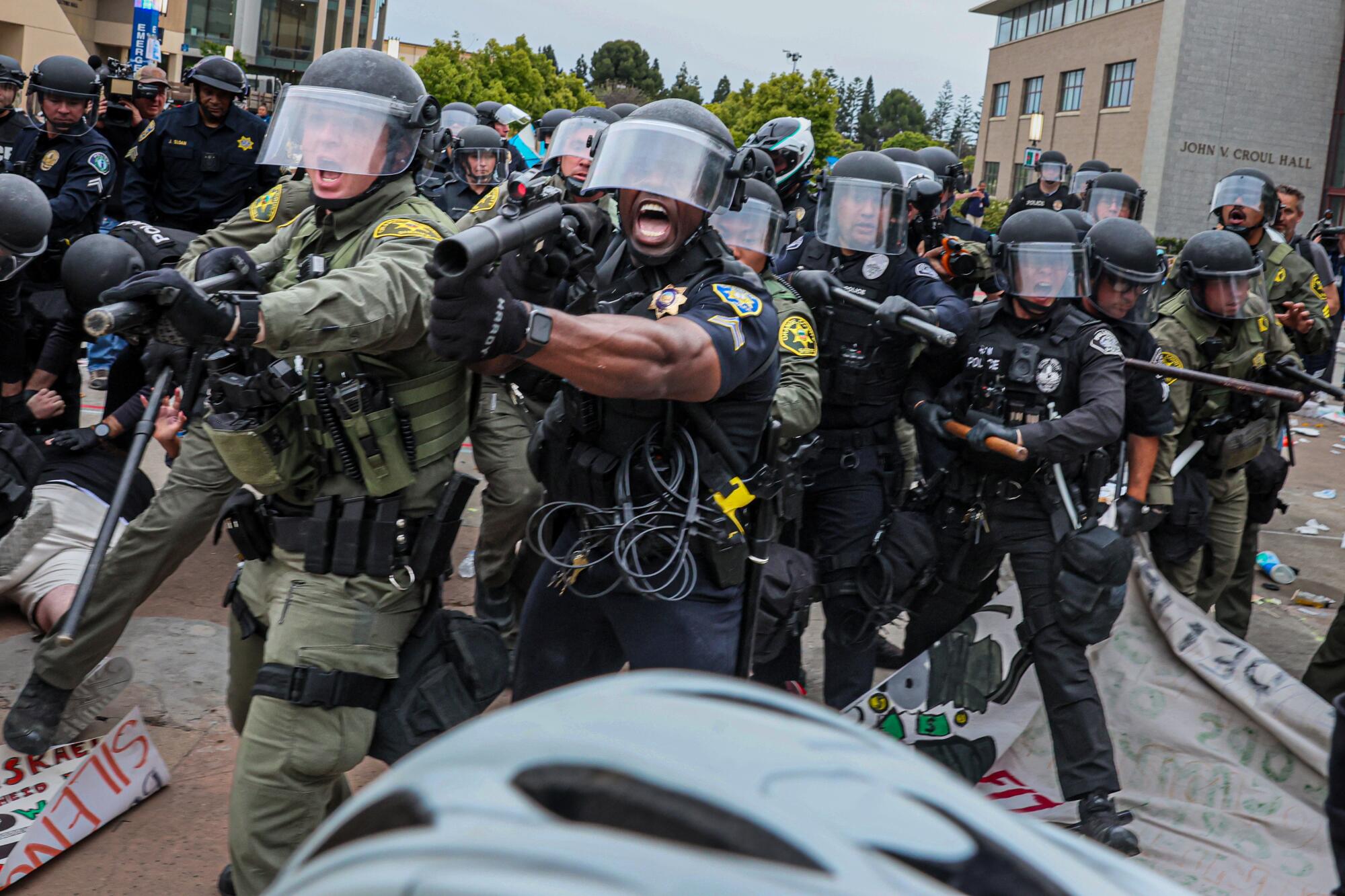 Officers in riot gear wielding various weapons and batons wade into a group of demonstrators.