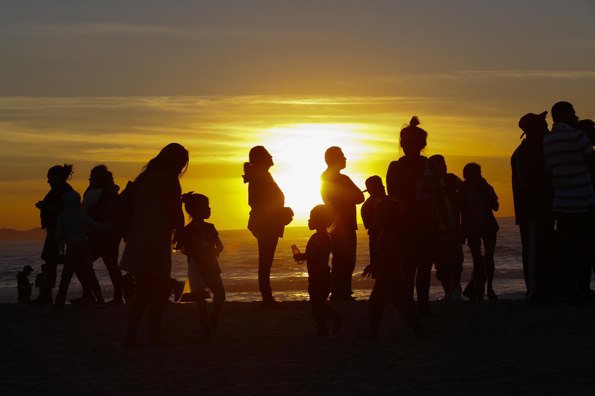 A group of Central American migrants silhouetted by the sunset at the beach by the border fence