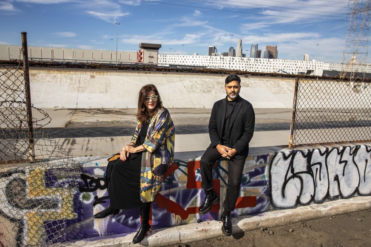 Mia Lehrer and Kush Parekh sit on a concrete rail on the banks of the L.A. River, downtown behind them