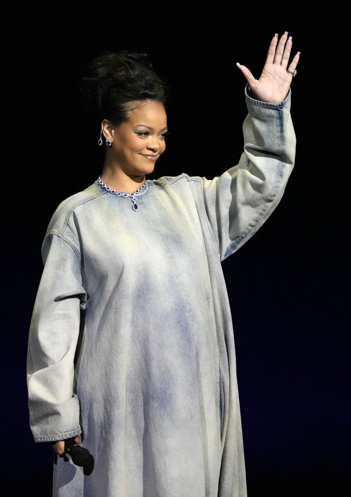 Rihanna holds up her left hand while standing on a stage in a long jean dress