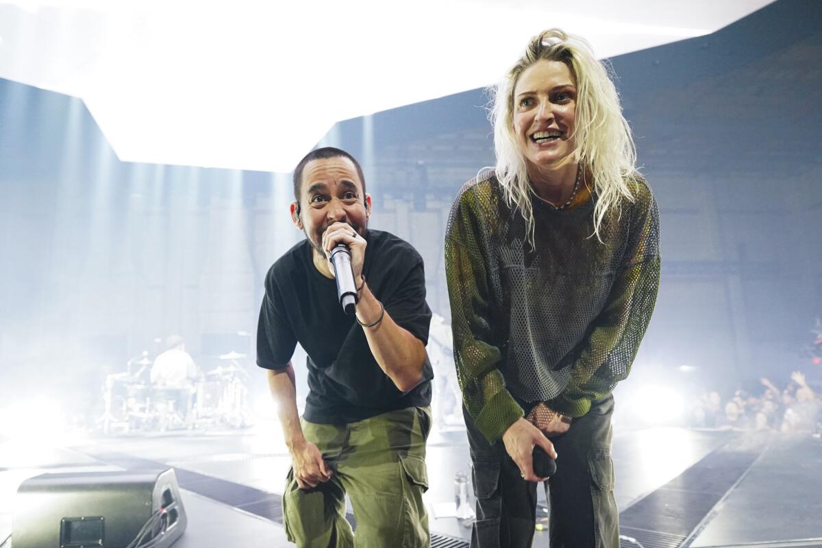 A man bending and singing onstage into a microphone next to a woman bending over while holding a microphone