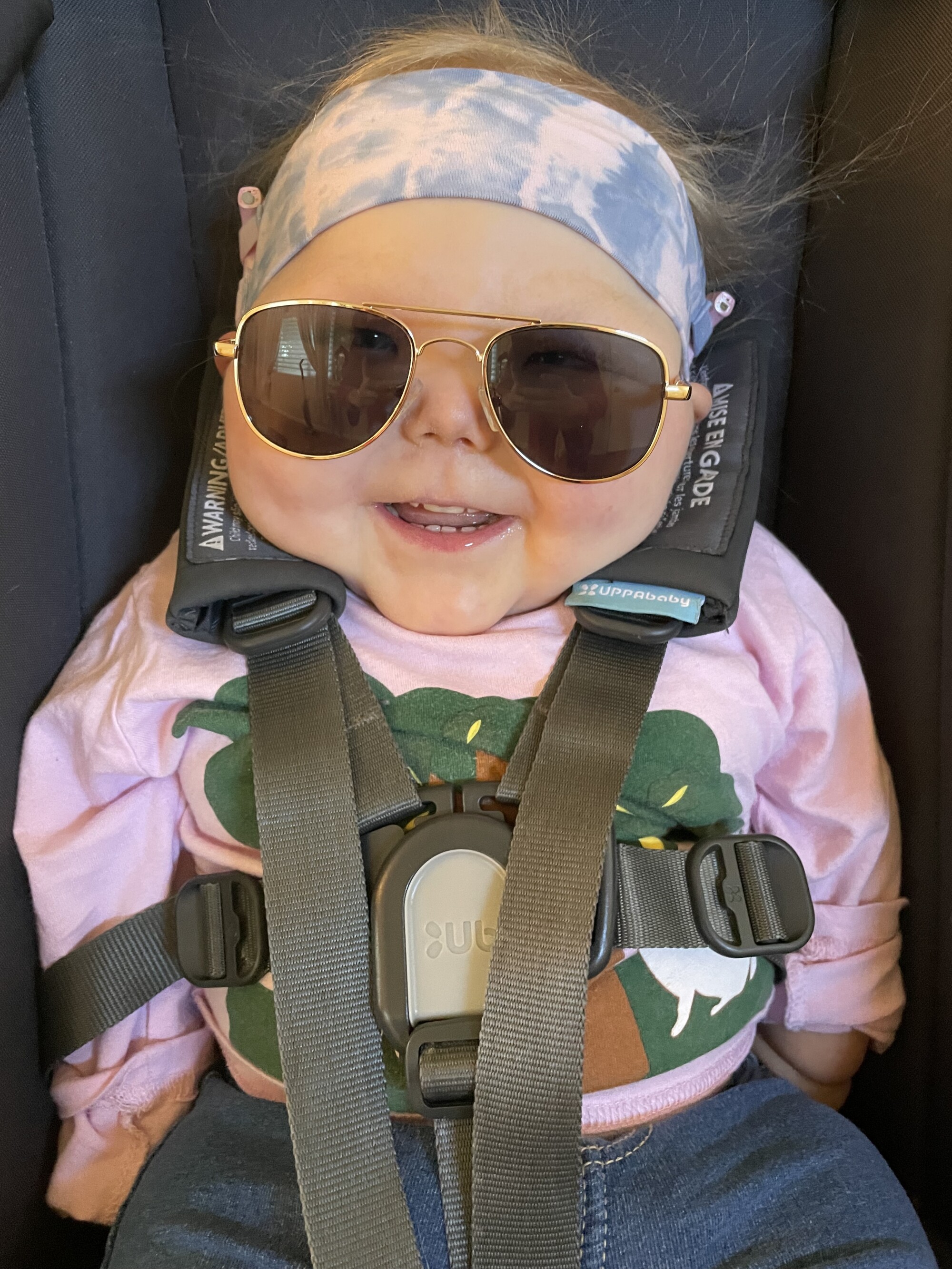 A smiling baby, wearing sunglasses and a headband, strapped into a car seat.