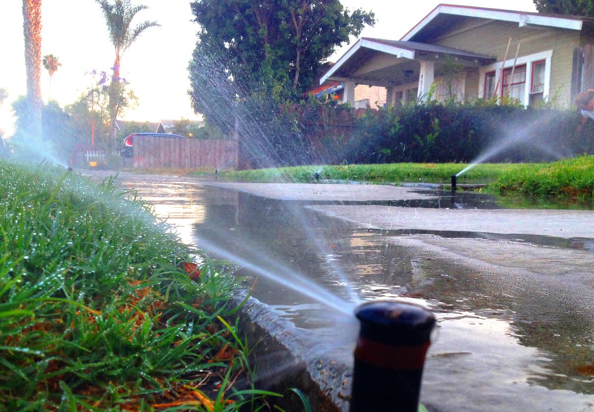 A ground-level view of sprinklers watering lawns and a sidewalk outside a single-family home.
