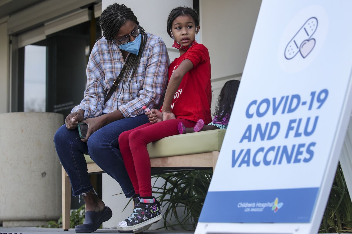 A woman sits beside a young girl next to a sign that reads "COVID-19 and flu vaccines."