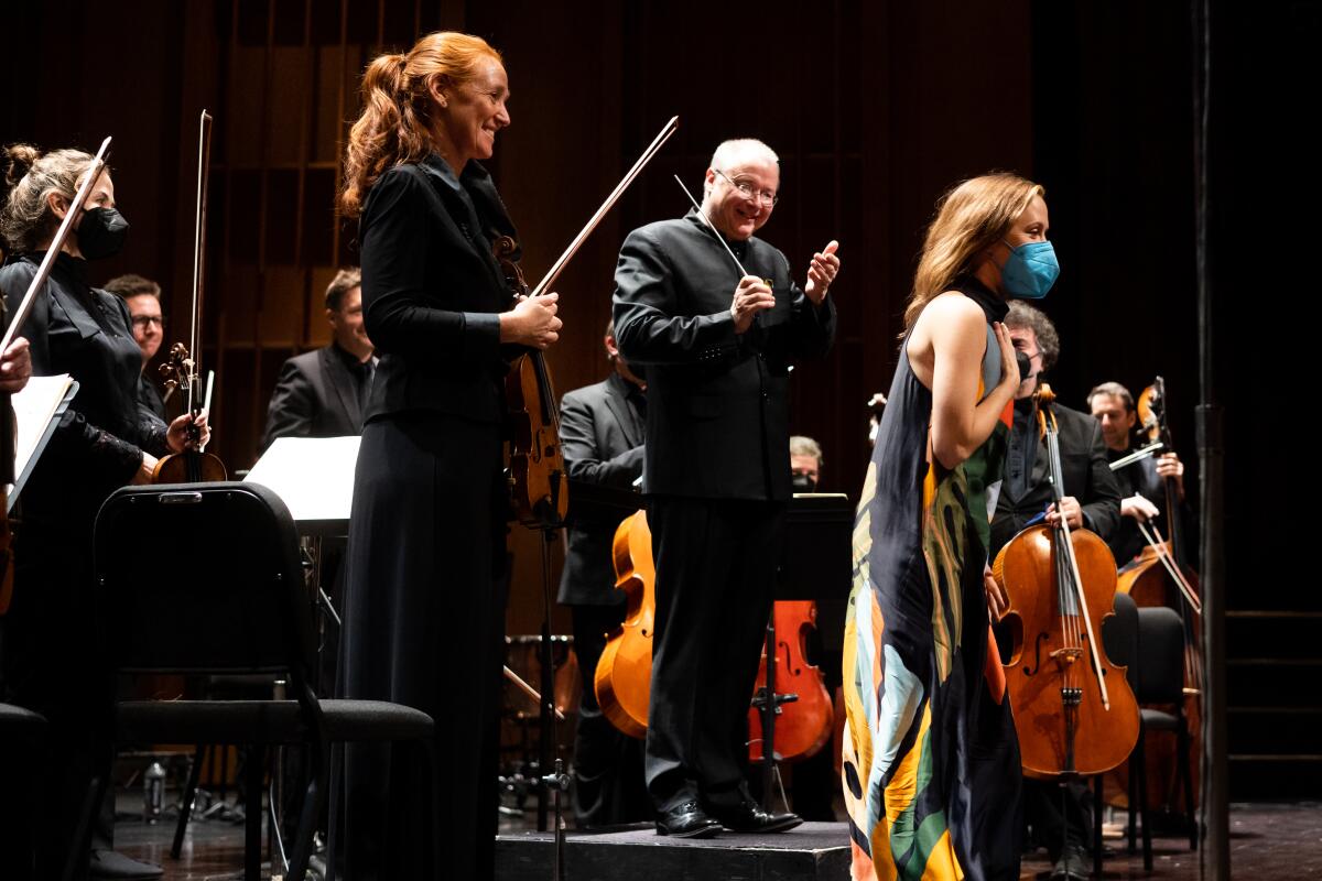 A woman in a long gown and mask bows. Behind, a conductor and musicians stand and smile.