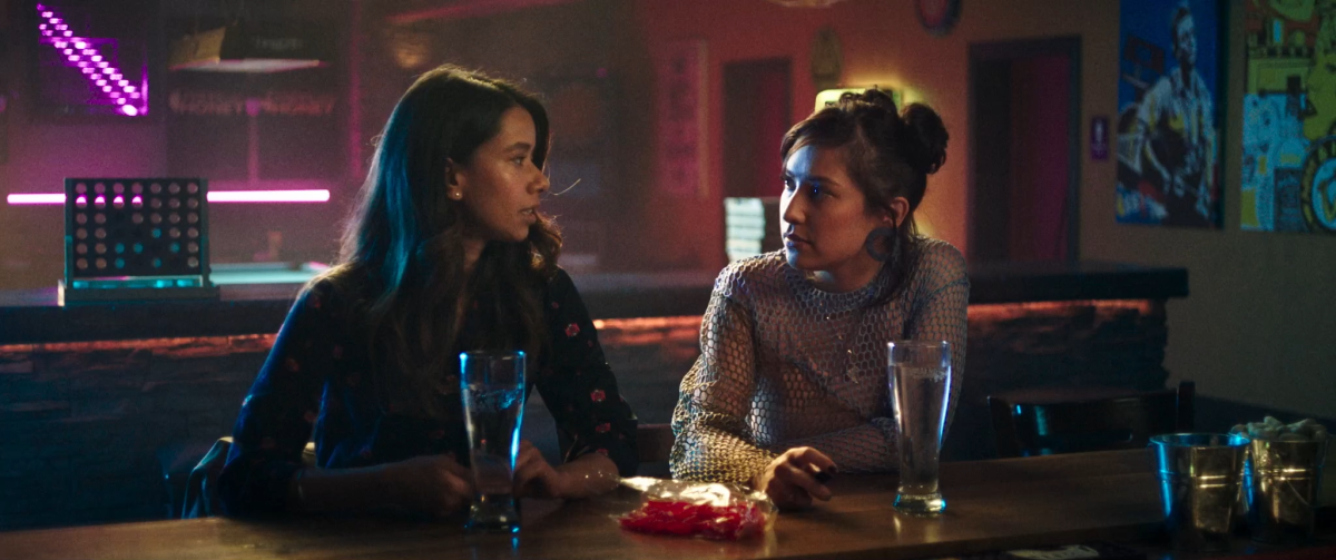 Two women sit together at a table with drinks in front of them in a scene from the film "Definition Please."