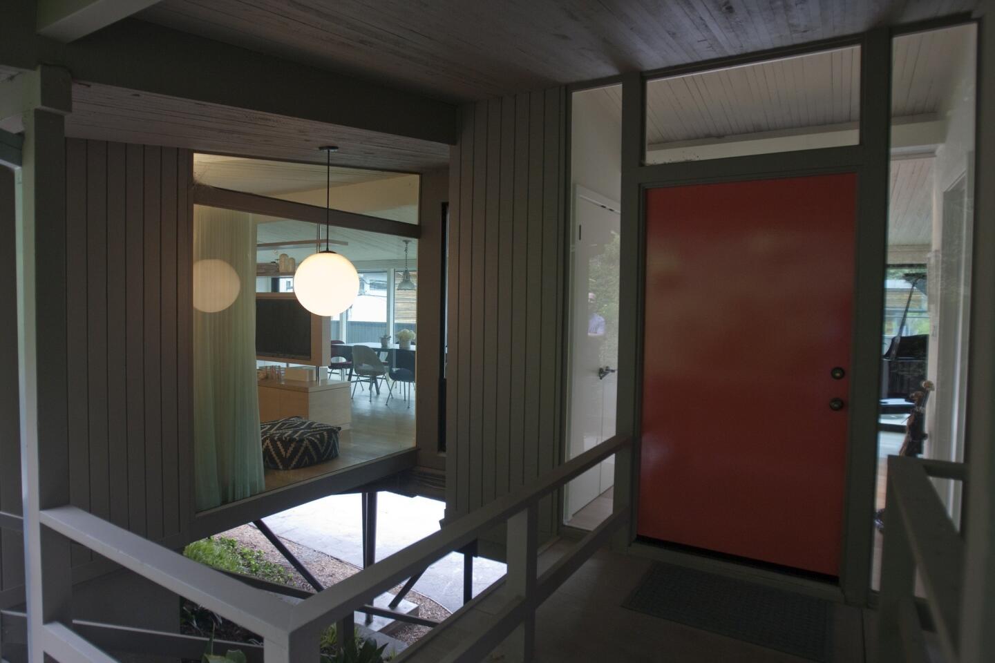 The front door, painted a bright orange inspired by Dutch painter Piet Mondrian, is original to the house. At lower left are some of the steel posts that elevate the house, giving it the feel of a Modernist treehouse.