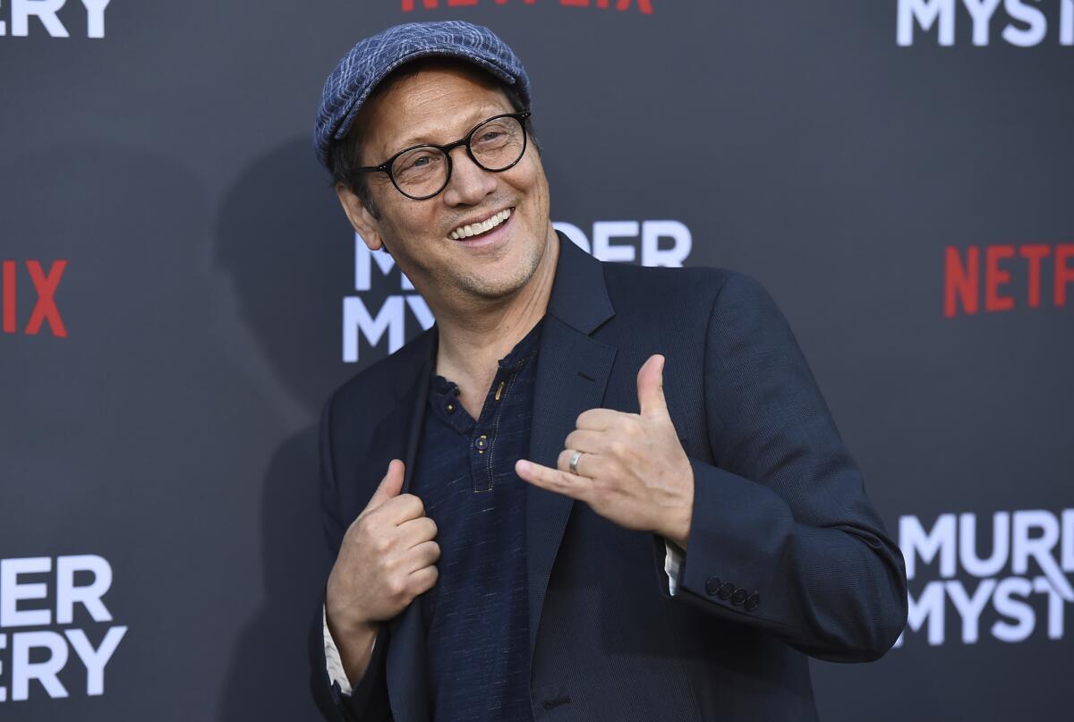Rob Schneider wears a dark suit as he flashes a shaka sign at a red carpet.