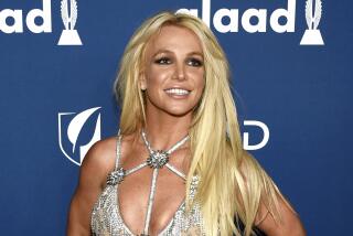 Britney Spears smiling in a strappy, bejeweled silver bra top against a blue backdrop