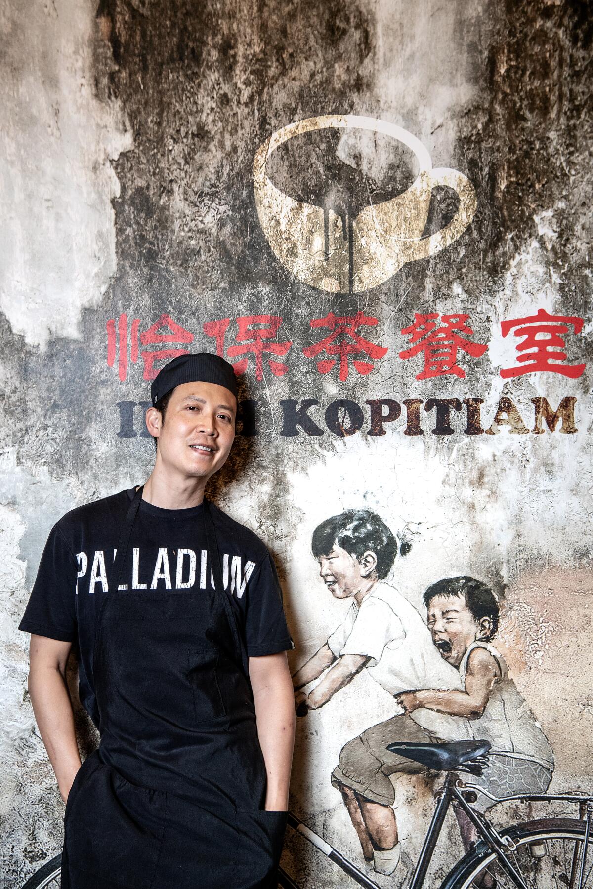 Kenji Tang stands beside a mural depicting two children on a bicycle.