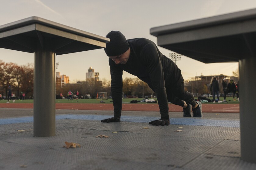 A man dressed in black workout clothes and gloves does a plank at a park, with a soccer game played in the distance.
