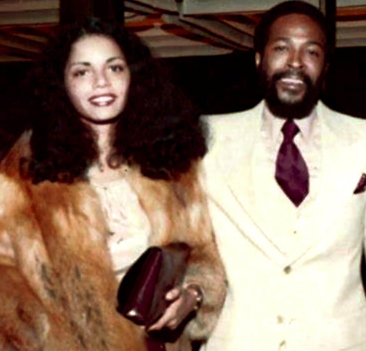 A woman in a fur coat stands with a man in a cream-colored suit